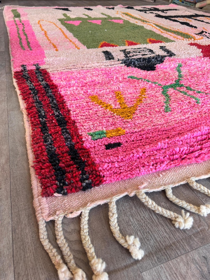 May include: A rectangular area rug featuring a high-pile texture with a vibrant pink base. The design includes abstract geometric shapes in green, black, yellow, and red, with thick black and red striped borders and cream-colored braided fringe.