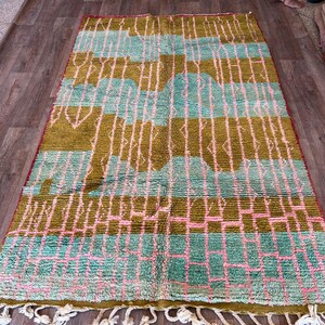 May include: A rectangular rug with a geometric pattern in shades of olive green, teal, and pink. The rug has a fringe along the bottom edge. The design includes vertical and horizontal lines, creating a grid-like appearance. The rug is placed on a wooden floor.
