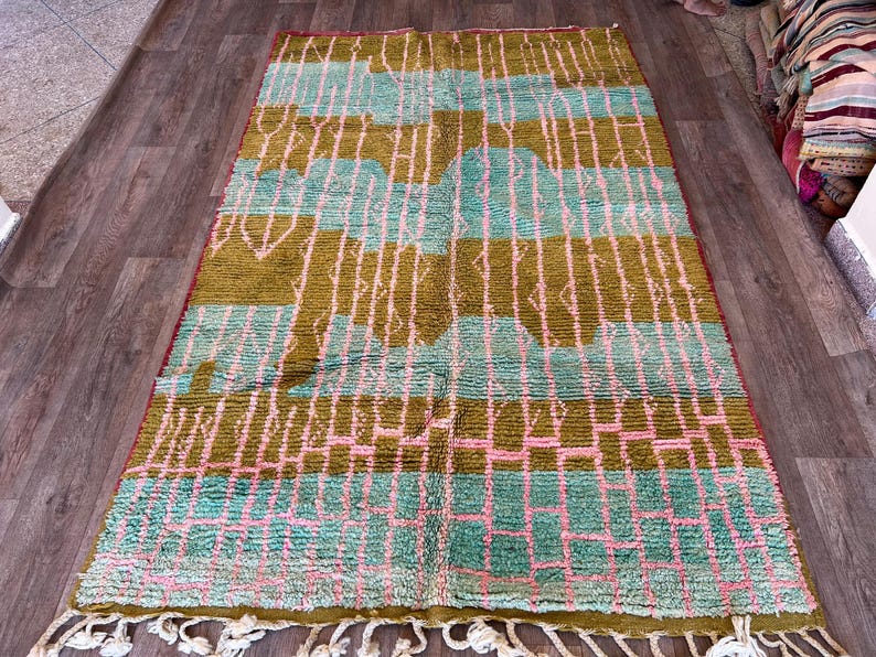 May include: A rectangular rug with a geometric pattern in shades of olive green, teal, and pink. The rug has a fringe along the bottom edge. The design includes vertical and horizontal lines, creating a grid-like appearance. The rug is placed on a wooden floor.