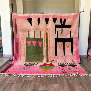 May include: A large rectangular area rug featuring a light pink background with a variety of abstract geometric shapes in black, olive green, bright pink, and orange. The rug includes a knotted fringe along the bottom edge and a thick border.