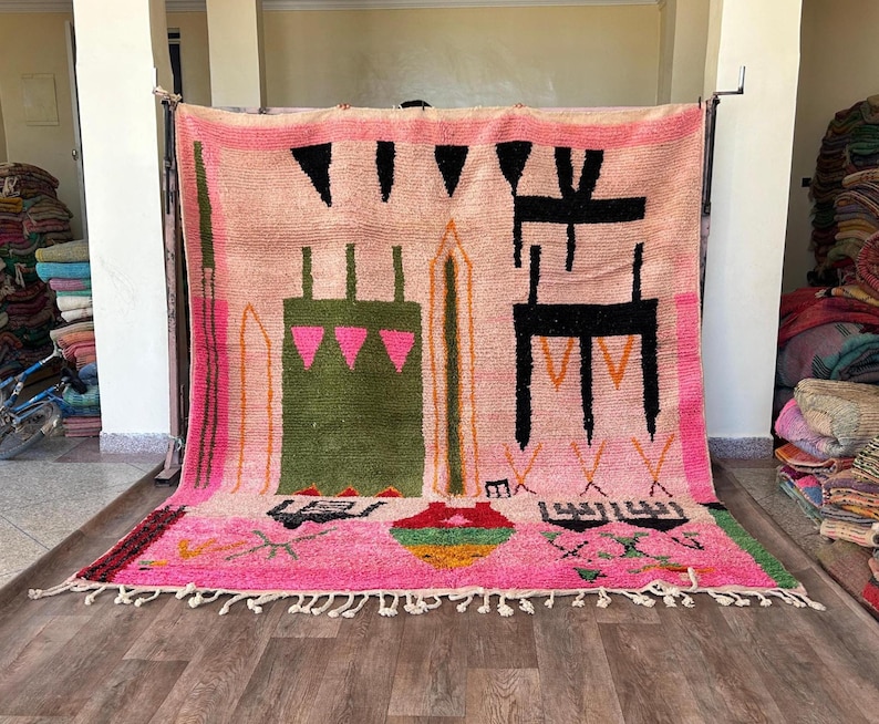 May include: A large rectangular area rug featuring a light pink background with a variety of abstract geometric shapes in black, olive green, bright pink, and orange. The rug includes a knotted fringe along the bottom edge and a thick border.