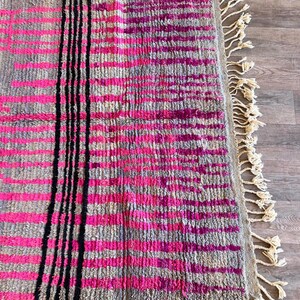 May include: A rectangular rug with a grey background and a pattern of horizontal pink stripes. Vertical black stripes run down the centre. The rug has a fringe of cream-coloured tassels along one edge.
