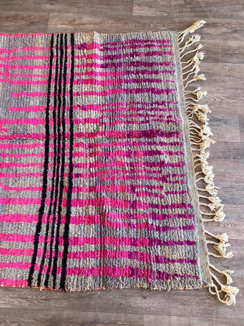 May include: A rectangular rug with a grey background and a pattern of horizontal pink stripes. Vertical black stripes run down the centre. The rug has a fringe of cream-coloured tassels along one edge.