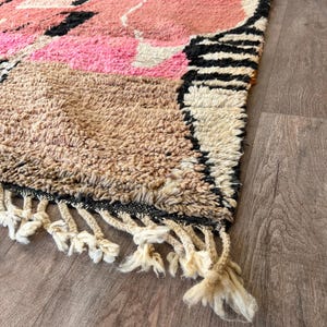 May include: A hand-woven rug with a geometric pattern in shades of pink, brown, black, and white. The rug has a fringe border.