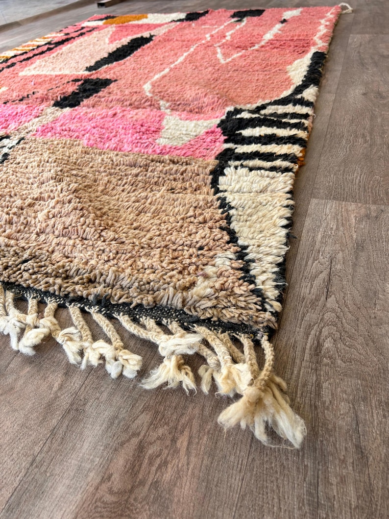 May include: A hand-woven rug with a geometric pattern in shades of pink, brown, black, and white. The rug has a fringe border.