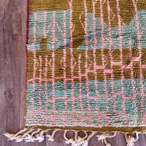 May include: A close-up of a textured rug with a geometric pattern. The rug features a combination of olive green, light blue, and pink colours. The rug has a fringe of white tassels along the bottom edge.