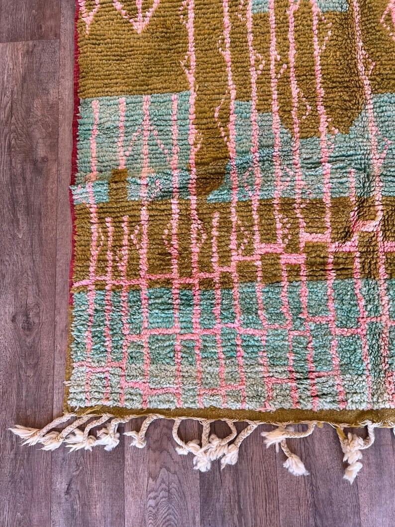 May include: A close-up of a textured rug with a geometric pattern. The rug features a combination of olive green, light blue, and pink colours. The rug has a fringe of white tassels along the bottom edge.