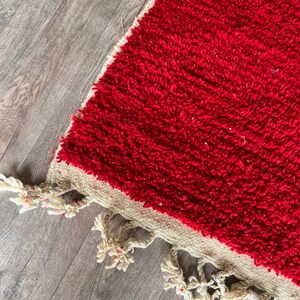 Peut inclure: Un tapis rectangulaire pr&eacute;sentant une texture &agrave; poils longs d'un rouge profond qui recouvre la majeure partie de la surface. Le tapis poss&egrave;de une bordure tiss&eacute;e beige naturel sur le bord, finie avec des pompons &agrave; franges blanc cass&eacute;.