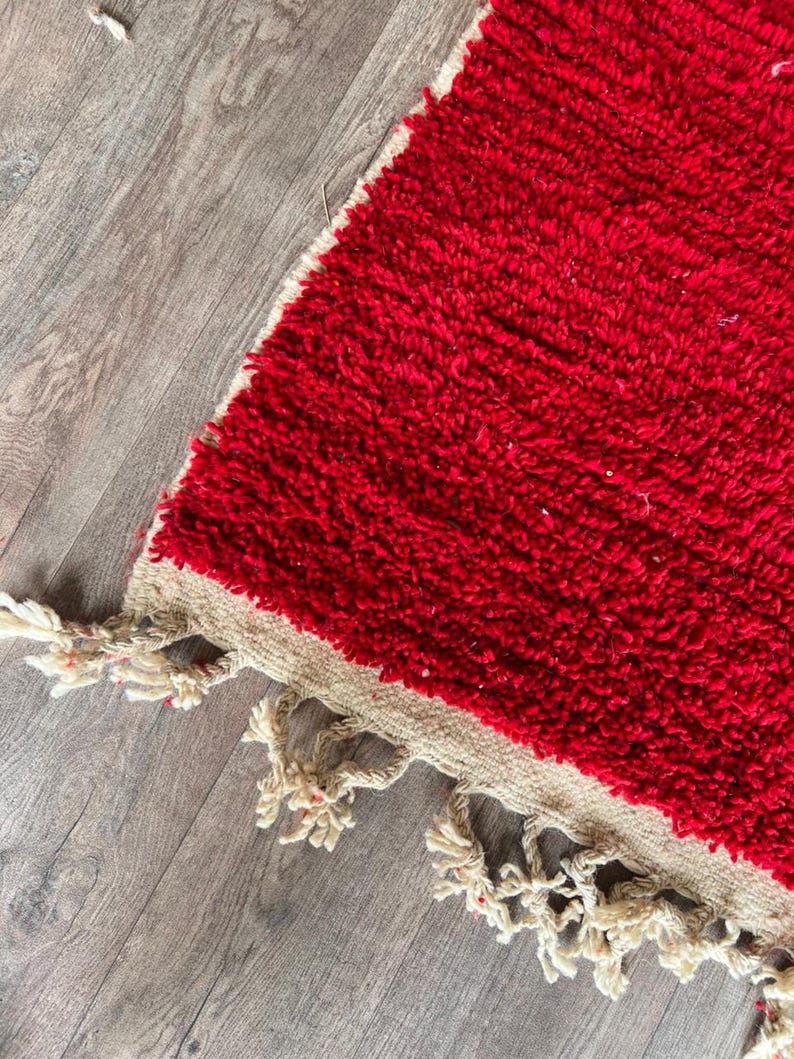 Peut inclure: Un tapis rectangulaire pr&eacute;sentant une texture &agrave; poils longs d'un rouge profond qui recouvre la majeure partie de la surface. Le tapis poss&egrave;de une bordure tiss&eacute;e beige naturel sur le bord, finie avec des pompons &agrave; franges blanc cass&eacute;.