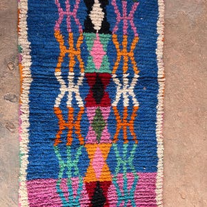 May include: A rectangular, hand-woven rug with a vibrant geometric pattern. The rug features a blue central field with pink, orange, green, and white diamond and cross-like designs. The edges are finished with a white border. The rug's texture appears soft and plush.
