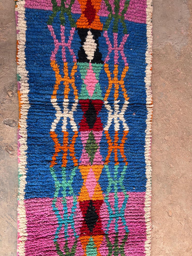 May include: A rectangular, hand-woven rug with a vibrant geometric pattern. The rug features a blue central field with pink, orange, green, and white diamond and cross-like designs. The edges are finished with a white border. The rug's texture appears soft and plush.