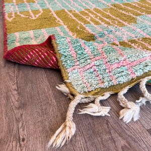 May include: A colourful, hand-woven rug with a geometric pattern. The rug features olive green, light blue, and pink hues. The edges are finished with tassels. The rug is folded over, revealing a red and pink patterned underside.