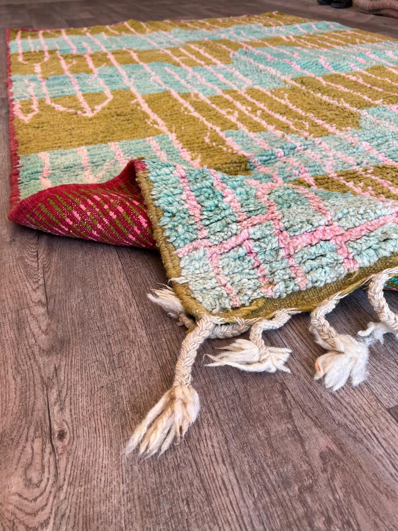 May include: A colourful, hand-woven rug with a geometric pattern. The rug features olive green, light blue, and pink hues. The edges are finished with tassels. The rug is folded over, revealing a red and pink patterned underside.