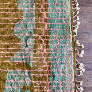 May include: A rectangular rug with a geometric pattern in olive green, light blue, and pink. The rug has a textured surface and a fringe border with cream-coloured tassels. The design features a grid-like pattern.