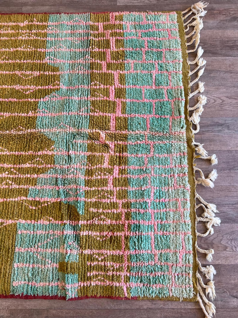 May include: A rectangular rug with a geometric pattern in olive green, light blue, and pink. The rug has a textured surface and a fringe border with cream-coloured tassels. The design features a grid-like pattern.
