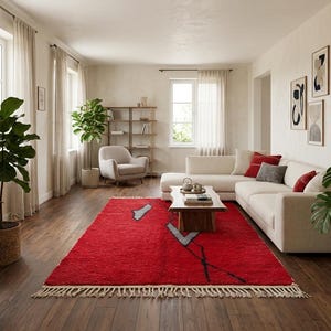 Peut inclure: Un tapis rectangulaire au fond rouge vif orn&eacute; de lignes g&eacute;om&eacute;triques abstraites noires et grises, avec des franges sur les bords. Le tapis est pos&eacute; sur un parquet en bois fonc&eacute; dans un salon avec un canap&eacute; blanc et une table basse en bois.
