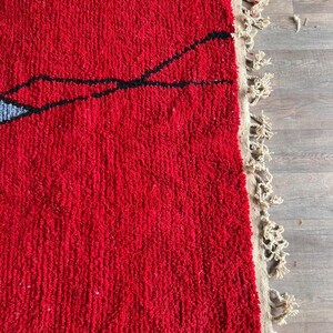 Peut inclure: Un tapis &agrave; poils longs et &eacute;pais pr&eacute;sentant une base rouge vif avec de fines lignes noires nou&eacute;es &agrave; la main formant un motif abstrait. Une petite section bleu clair est visible sur le bord gauche. Le tapis a une frange beige sur le c&ocirc;t&eacute; droit.