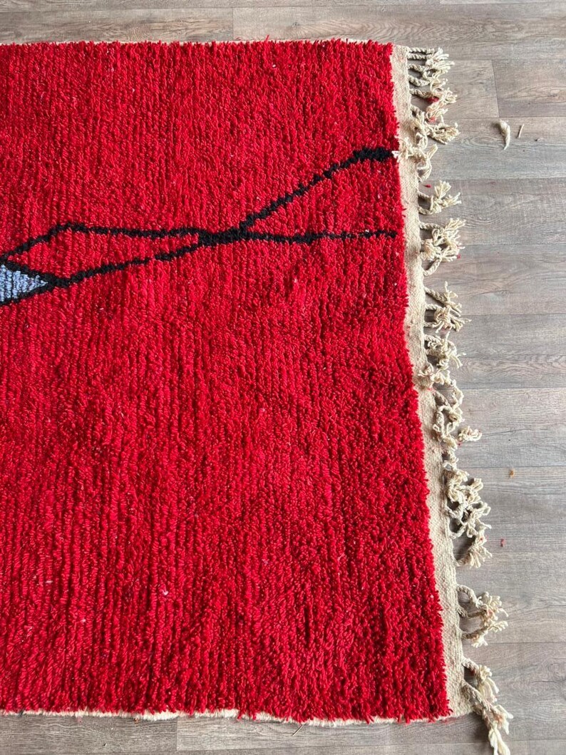 Peut inclure: Un tapis &agrave; poils longs et &eacute;pais pr&eacute;sentant une base rouge vif avec de fines lignes noires nou&eacute;es &agrave; la main formant un motif abstrait. Une petite section bleu clair est visible sur le bord gauche. Le tapis a une frange beige sur le c&ocirc;t&eacute; droit.