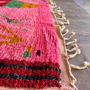 May include: A hand-knotted wool rug featuring a vibrant pink base with geometric patterns in green, yellow, and black. The rug has a thick, textured pile and is finished with long, cream-colored braided fringe tassels along the side edge.
