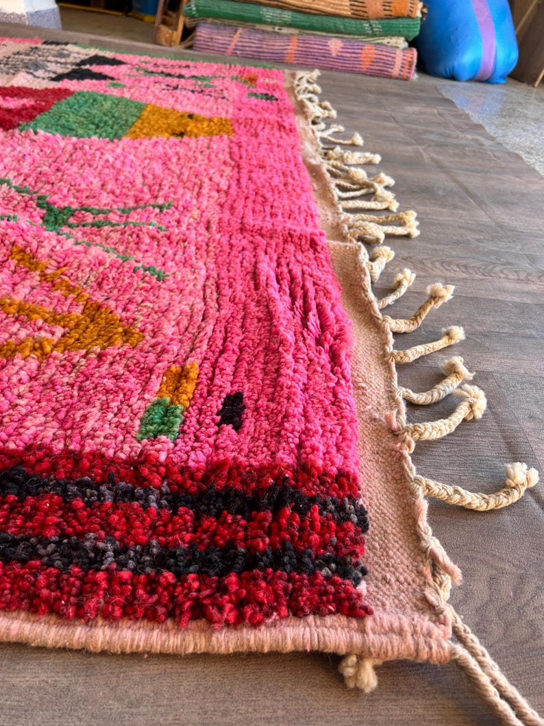 May include: A hand-knotted wool rug featuring a vibrant pink base with geometric patterns in green, yellow, and black. The rug has a thick, textured pile and is finished with long, cream-colored braided fringe tassels along the side edge.