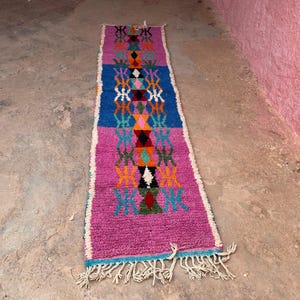 May include: A rectangular, hand-woven rug with a vibrant design. The rug features a central pattern of diamonds and geometric shapes in shades of pink, blue, orange, and green. The edges are trimmed with white fringe. The rug is approximately 152 cm long.