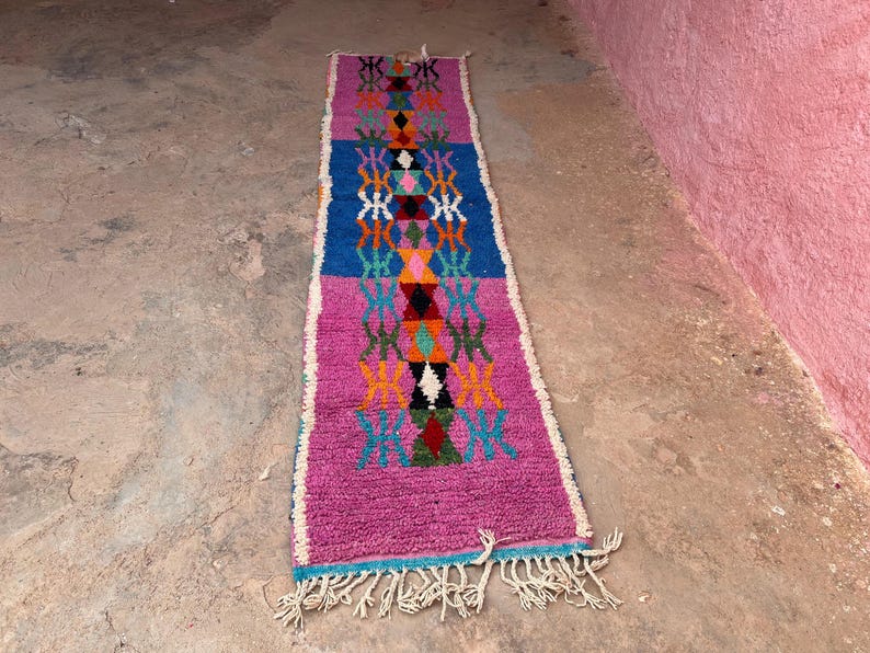 May include: A rectangular, hand-woven rug with a vibrant design. The rug features a central pattern of diamonds and geometric shapes in shades of pink, blue, orange, and green. The edges are trimmed with white fringe. The rug is approximately 152 cm long.