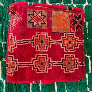 May include: A square, red Moroccan pouf with geometric patterns in orange, black, and gray. The pouf sits on a green and white textured rug. The pouf's design includes star and cross-like shapes.