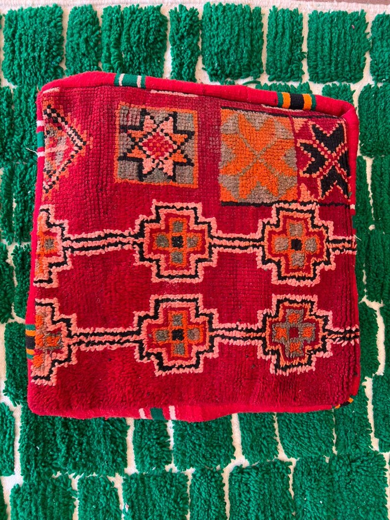 May include: A square, red Moroccan pouf with geometric patterns in orange, black, and gray. The pouf sits on a green and white textured rug. The pouf's design includes star and cross-like shapes.