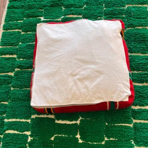 May include: A square, red floor cushion with a white top, resting on a green and white patterned rug. The cushion has a striped border in red, yellow, and green. The rug has a geometric design.