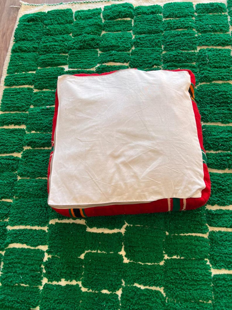 May include: A square, red floor cushion with a white top, resting on a green and white patterned rug. The cushion has a striped border in red, yellow, and green. The rug has a geometric design.