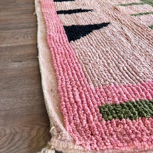 May include: A thick, textured rug featuring a light pink base with a prominent pink border. The surface displays geometric patterns in black, green, and orange. The edge of the rug is finished with a braided fringe detail at the corner.