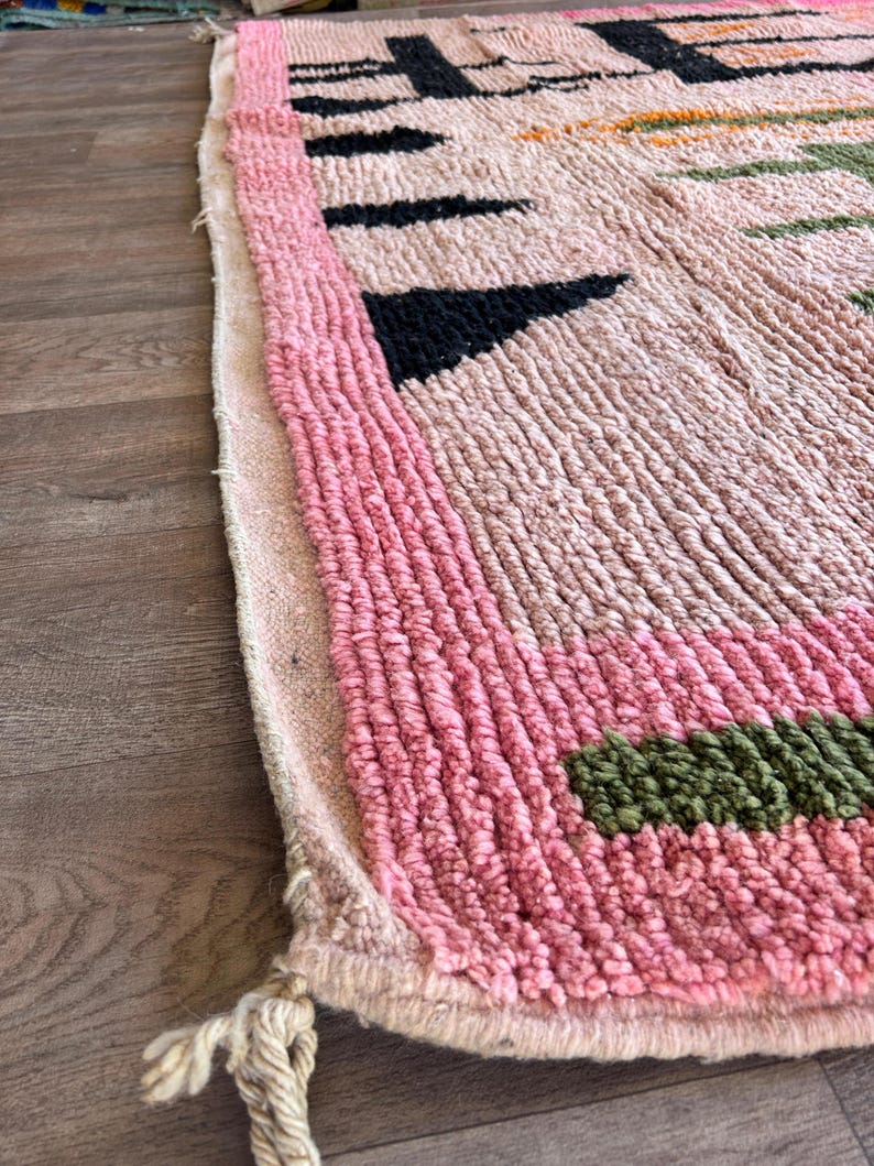 May include: A thick, textured rug featuring a light pink base with a prominent pink border. The surface displays geometric patterns in black, green, and orange. The edge of the rug is finished with a braided fringe detail at the corner.
