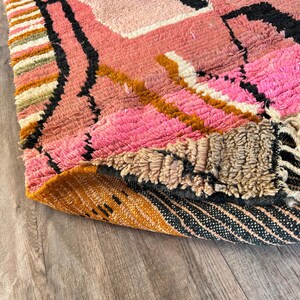 May include: Close-up of a pink, orange, and black patterned rug with a fringe edge. The rug is folded over on itself, revealing the underside of the rug, which is a brown and black striped pattern.