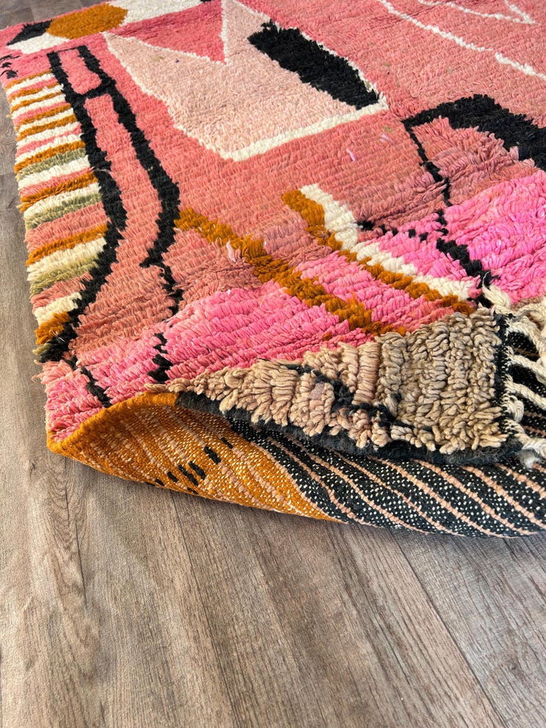 May include: Close-up of a pink, orange, and black patterned rug with a fringe edge. The rug is folded over on itself, revealing the underside of the rug, which is a brown and black striped pattern.