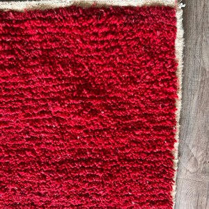 Peut inclure: Un tapis rectangulaire pr&eacute;sentant une texture &agrave; poils longs d'un rouge profond. Le tapis poss&egrave;de une bordure de couleur beige naturel sur les bords sup&eacute;rieur et droit, avec des fibres l&acirc;ches visibles aux coins du tissage.