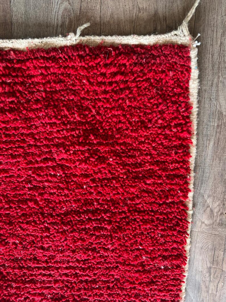 Peut inclure: Un tapis rectangulaire pr&eacute;sentant une texture &agrave; poils longs d'un rouge profond. Le tapis poss&egrave;de une bordure de couleur beige naturel sur les bords sup&eacute;rieur et droit, avec des fibres l&acirc;ches visibles aux coins du tissage.