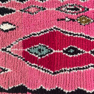 Puede incluir: Primer plano de una alfombra marroqu&iacute; rosa y roja con un patr&oacute;n de diamantes geom&eacute;tricos. La alfombra presenta un dise&ntilde;o texturizado y tejido con detalles en negro, blanco y turquesa. La franja de la alfombra es negra.
