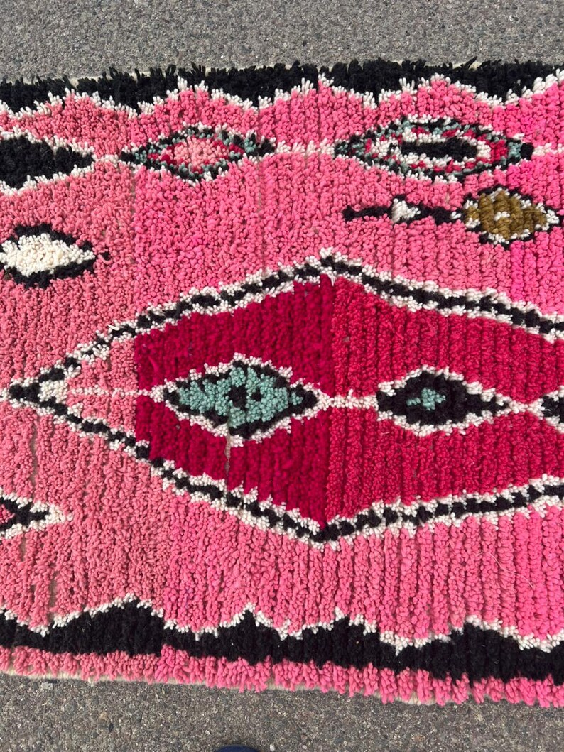 Puede incluir: Primer plano de una alfombra marroqu&iacute; rosa y roja con un patr&oacute;n de diamantes geom&eacute;tricos. La alfombra presenta un dise&ntilde;o texturizado y tejido con detalles en negro, blanco y turquesa. La franja de la alfombra es negra.