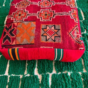 May include: A square, red floor cushion with a geometric pattern in orange, black, and gray. The cushion is placed on a green and white patterned rug. The cushion has a thick, textured appearance.