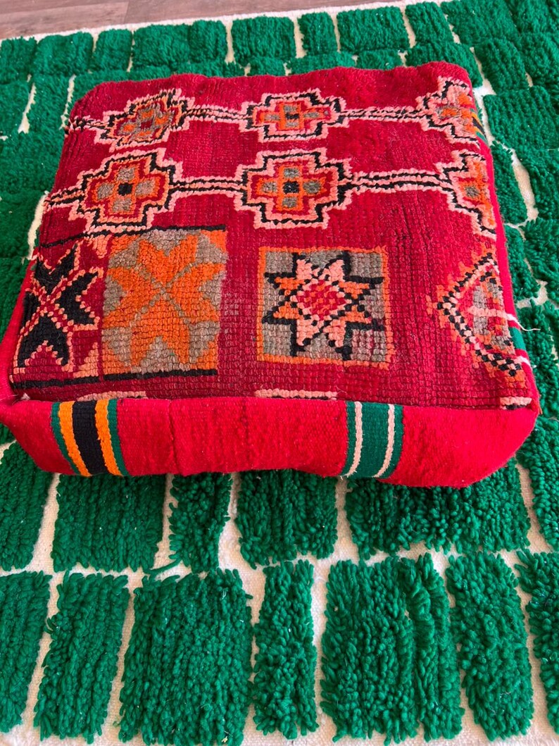 May include: A square, red floor cushion with a geometric pattern in orange, black, and gray. The cushion is placed on a green and white patterned rug. The cushion has a thick, textured appearance.