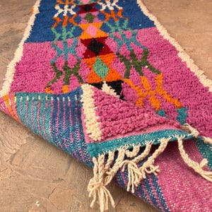 May include: A vibrant, hand-woven runner rug with a geometric design. The rug features a palette of pink, blue, orange, and green, with white fringe. The rug is partially rolled, showing its textured weave. Measures approximately 200cm in length.