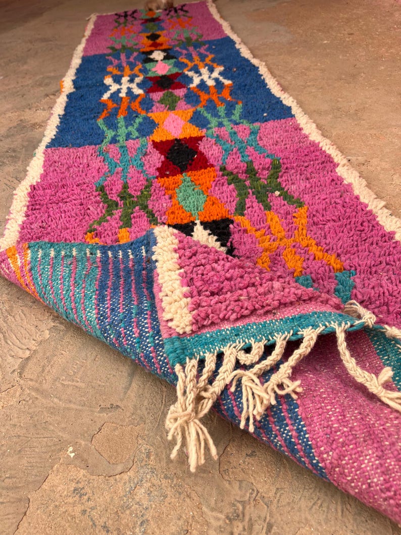 May include: A vibrant, hand-woven runner rug with a geometric design. The rug features a palette of pink, blue, orange, and green, with white fringe. The rug is partially rolled, showing its textured weave. Measures approximately 200cm in length.