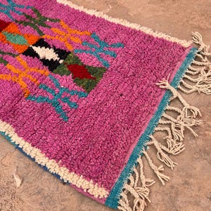 May include: A colourful, hand-woven rug with a textured surface. The rug has a bright pink field with geometric patterns in orange, teal, black, and white. Cream-coloured fringe edges the rug. The rug's dimensions are not visible.