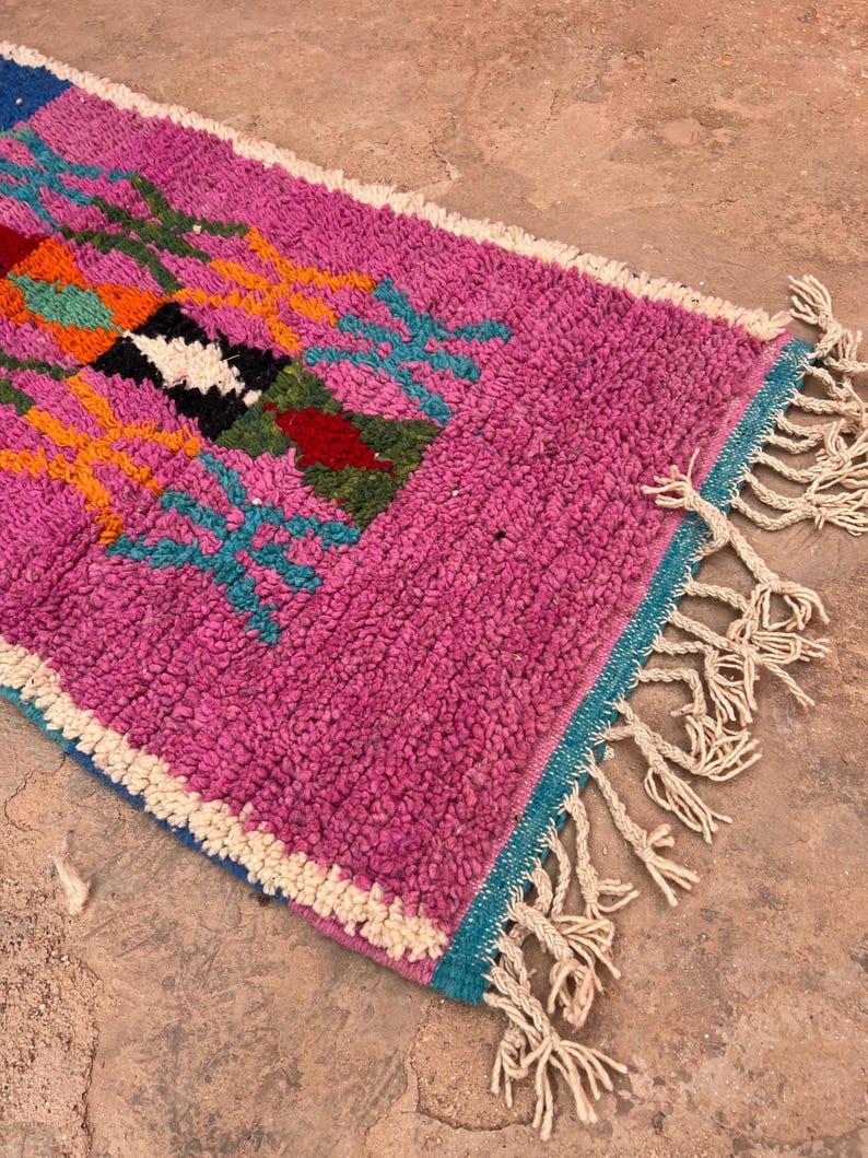 May include: A colourful, hand-woven rug with a textured surface. The rug has a bright pink field with geometric patterns in orange, teal, black, and white. Cream-coloured fringe edges the rug. The rug's dimensions are not visible.