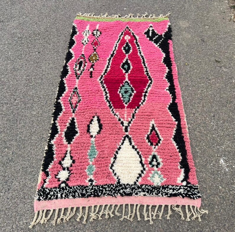 Puede incluir: Una alfombra marroqu&iacute; rosa con un dise&ntilde;o geom&eacute;trico. La alfombra presenta una forma de diamante central en rojo y negro, rodeada de formas de diamante y rombo m&aacute;s peque&ntilde;as en blanco, negro y verde azulado. La alfombra tiene flecos en la parte inferior y un borde verde.