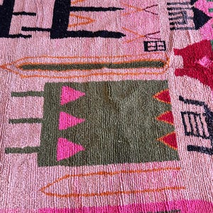 May include: A textured rug with a light pink base featuring various geometric shapes and symbols in black, olive green, bright pink, and orange. The design includes abstract rectangular blocks, triangular patterns, and linear motifs.