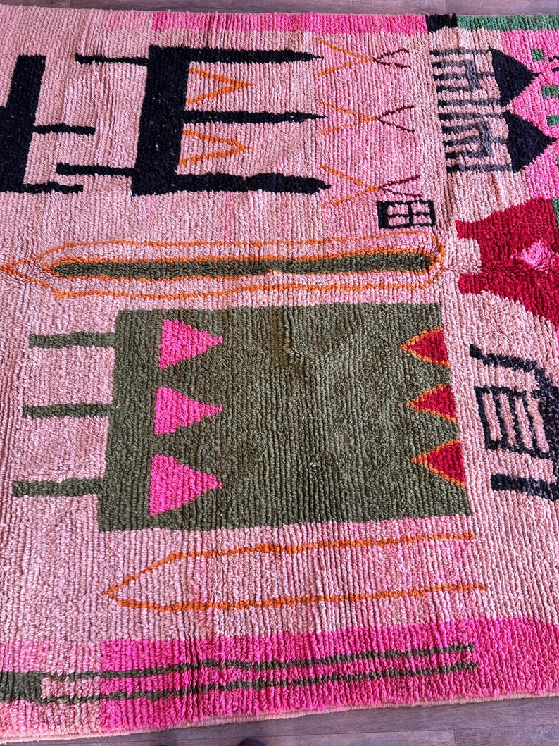 May include: A textured rug with a light pink base featuring various geometric shapes and symbols in black, olive green, bright pink, and orange. The design includes abstract rectangular blocks, triangular patterns, and linear motifs.