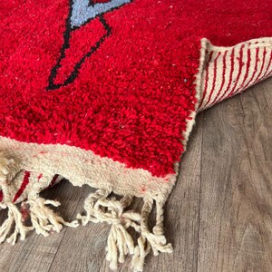 Peut inclure: Un tapis &agrave; poils longs rouge &eacute;pais avec un motif graphique abstrait noir et bleu clair. Le tapis pr&eacute;sente une bordure cr&egrave;me avec des pompons &agrave; franges &agrave; l'extr&eacute;mit&eacute;. Le dessous du tapis est visible, montrant un motif tiss&eacute; &agrave; rayures cr&egrave;me et rouge.