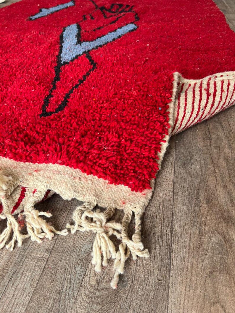 Peut inclure: Un tapis &agrave; poils longs rouge &eacute;pais avec un motif graphique abstrait noir et bleu clair. Le tapis pr&eacute;sente une bordure cr&egrave;me avec des pompons &agrave; franges &agrave; l'extr&eacute;mit&eacute;. Le dessous du tapis est visible, montrant un motif tiss&eacute; &agrave; rayures cr&egrave;me et rouge.