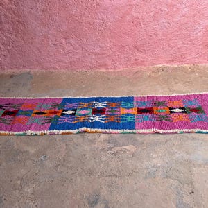 May include: A rectangular, hand-woven rug with a vibrant geometric pattern. The rug features a central blue section flanked by pink borders. The design incorporates orange, green, and white accents, with fringed edges.
